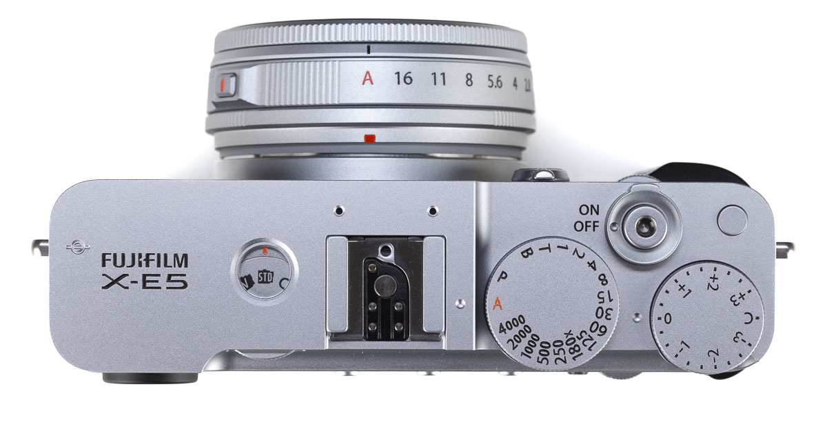 Sulla calotta della Fujifilm X-E5 campeggiano la ghiera dei tempi, il correttore dell'esposizione, il pulsante di scatto coassiale alla leva d'accensione, un tasto Fn "muto" e personalizzabile e il caratteristico oblò attraverso il quale osservare la simulazione pellicola utilizzata per le riprese fotografiche e video.
