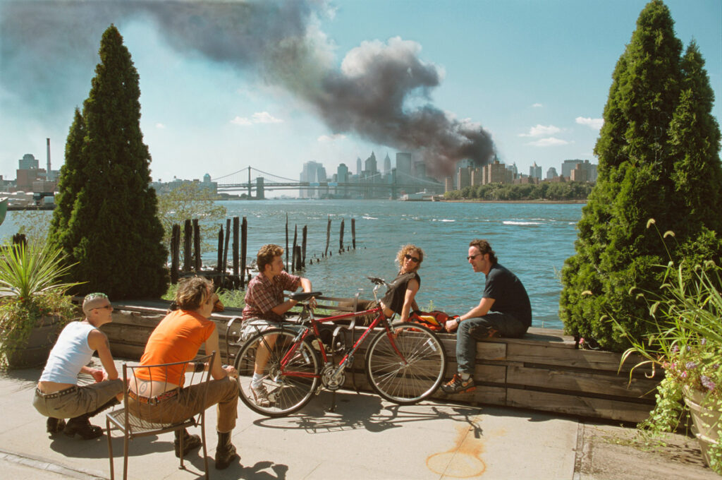 Giovani in pausa pranzo lungo l'East River mentre un'enorme colonna di fumo si alza da Lower Manhattan dopo l'attacco al World Trade Center, New York City, l'11 settembre 2001. © Thomas Hoepker/Magnum Photos